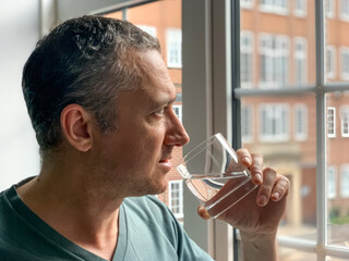 Man is drinking water from glass, staying by the window in Apartment. Middle aged man is drinking water to stay hydrated Thirsty man having hangover Pure life still water - obrazy, fototapety, plakaty