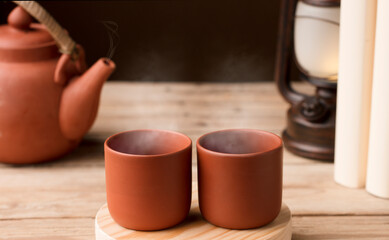 Two cups with hot steamed brown clay tea or coffee, on a wooden table with partial view of the lamp...