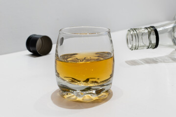 Glass with whiskey on white table close-up on blurred background of empty bottle and cap