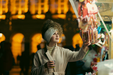 A portrait of a beautiful young woman holding a cup of mulled wine to warm up on the street with Christmas decorations.