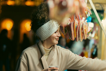 A portrait of a beautiful young woman holding a cup of mulled wine to warm up on the street with Christmas decorations.