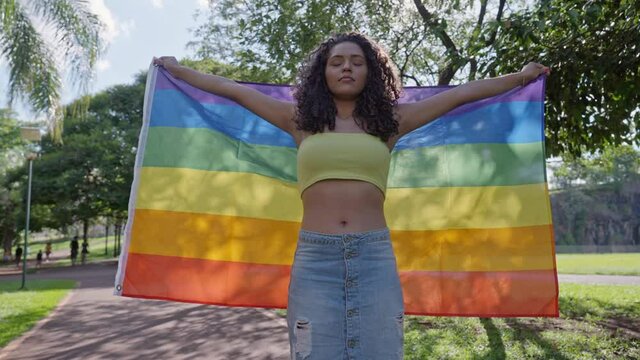 Young curly hair woman covering with lgbt pride flag. Alone. One. Keeping fist up, covering LGBT flag. LGBT+ flag on outdoor background. 4K.