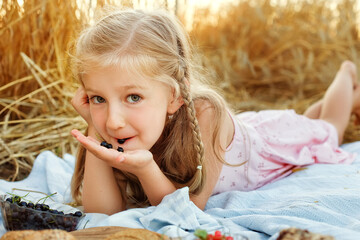 A cute blonde is lying on a blanket in a wheat field. Picnic in nature with milk, bread, berries. The girl is eating black currants. The child and the countryside.