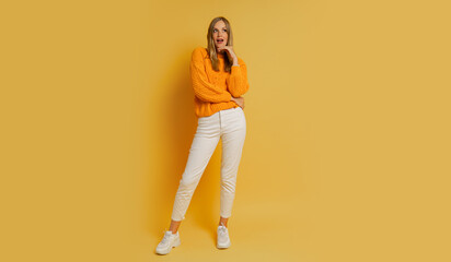 Fototapeta premium Blond woman with suprice face in orange stylish autumn sweater posing over yellow background in studio. Full lenght.