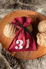Fototapeta na wymiar Red textile bag tied with green thread with date number thirty one, twine and natural rope coils on wooden round tray on faux fur blanket. Christmas home interior decor. Ball pet toy. Top view.