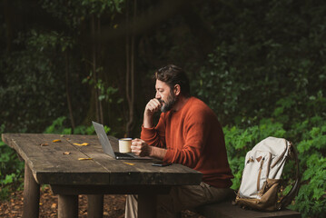 Mature worker man sit down on a wooden bench and work on the table in the middle of park outdoors forest woods alone. Backpack and laptop computer with people busy in modern job connection activity - Powered by Adobe