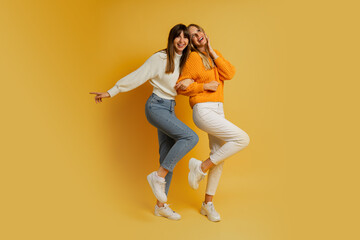 Fototapeta na wymiar Two pretty women , best friends in stylish autumn casual clothes having fun over yellow background in studio. Full lenght.
