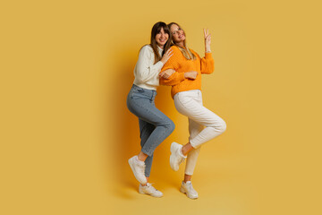 Fototapeta na wymiar Two pretty women , best friends in stylish autumn casual clothes having fun over yellow background in studio. Full lenght.