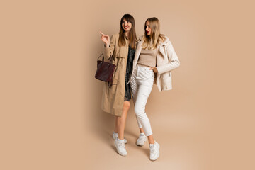 Two elegant women in stylish winter outfit posing over beige background. Wearing coat and jacket....