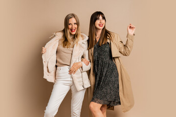 Two elegant women in stylish winter outfit posing over beige background. Wearing coat and jacket....