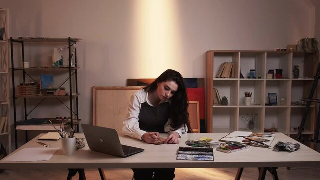 Art school. Female artist. Tutorial video. Pretty woman making draft on paper looking laptop in light shadow room interior.
