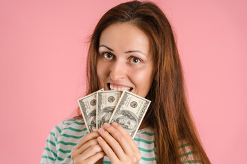 Portrait of a beautiful girl covering her face with dollar bills and pretending to bite money. Pink background