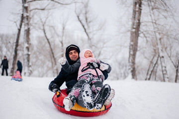 little kid smiling and sliding down hill on sled with father wearing warm winter clothes in forest. Astonishing background full of white color and snow. 