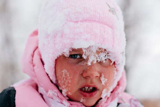 Closeup photo of female kid with sad face covered with snow and about to cry wearing pink winter clothes in forest. Astonishing background full of white color and snow. 
