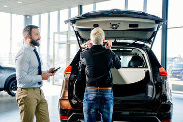 Adult blond caucasian businesswoman with a salesman examines the trunk of a new car in a car dealership.