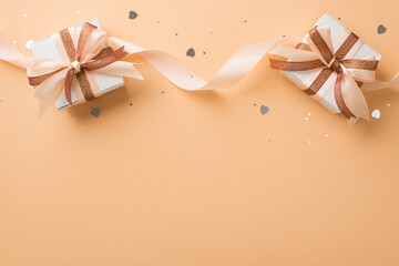Top view photo of valentine's day decorations white gift boxes with glowing brown and light beige curly ribbon bows heart shaped confetti and sequins on isolated beige background with empty space