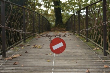 Typical white bar on red background no entry sign hanging crooked on a rusty steel chain cordoning off a rustic decorative iron bridge and the light woodland behind it