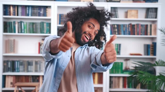Cheerful young Indian man office worker performs simple dance showing thumbs up making cool approve gesture ok yes in room with bookcase. Love for life and open expression of emotions concept.