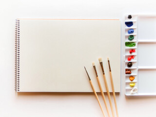 スケッチブックの白い画用紙と4本の絵筆と12色の絵の具を置いたプラスチックの水彩パレット。白背景で真上から撮影。