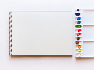 スケッチブックの白い画用紙と12色の絵の具を置いたプラスチックの水彩パレット。白背景で真上から撮影。