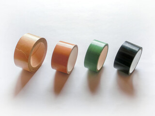 白い紙の上を転がる4種類のテープ。クラフトテープ、布テープ、養生テープ、黒布テープ。