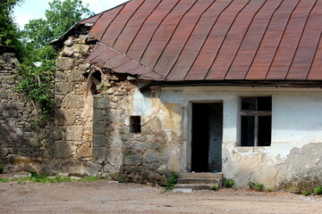 Fototapeta na wymiar Abandoned old traditional stone house with missing doors and broken windows covered with destroyed rusted metal roof tiles surrounded with gravel road and tall trees in background on warm sunny spring