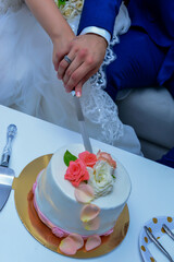 Obraz na płótnie Canvas bride and groom cut the wedding cake. The cake is decorated with beige and peach-colored roses. groom is dressed in blue wedding suit and bride in a white wedding dress..