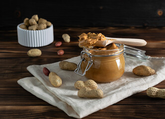 A wooden spoon with peanut paste lies on a glass jar. Raw, unpeeled peanuts in a white plaster bowl in the background. Nuts on a light napkin in the foreground