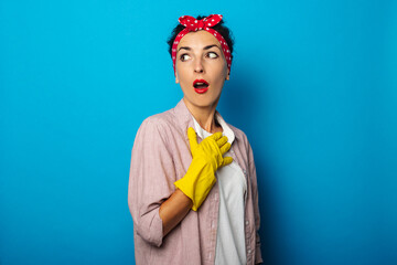 Cleaning. Surprised young woman with bandage on hair wearing cleaning gloves on blue background.