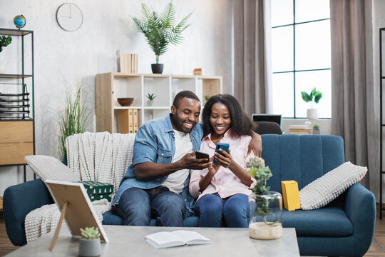 Happy couple of african american man and woman relaxing on cozy sofa with modern smartphones in hands. Two young people surfing internet during free time at home.