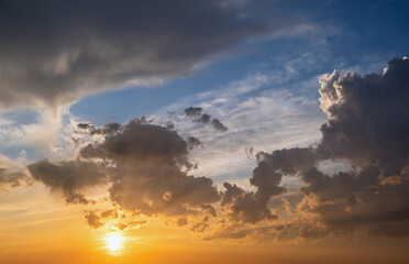 Dramatic sunset landscape with puffy clouds lit by orange setting sun and blue sky.