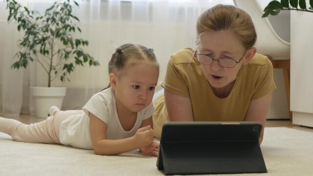 Grandmother reads e-book on tablet for granddaughter. Grandma and granddaughter spend time together. Concept of warm relationships between different aged relatives in family.
