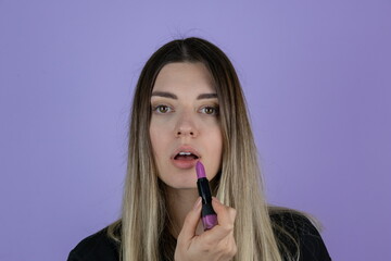 Close up shot of charming adorable lady with straight hair holding purple lipstick and applying it. Purple background. Copy and paste. Pampering, lips correction, dermatology, dream, healthcare.