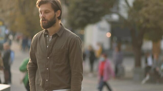 Attractive young bearded man with laptop waiting for somebody in the city street. Portrait of handsome man waiting. High quality 4k footage