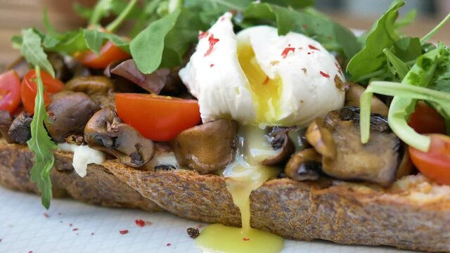 Fork and knife cut poached egg and yolk flows out on canape with mushrooms tomatoes and ruccola on piece of wholemeal bread macro