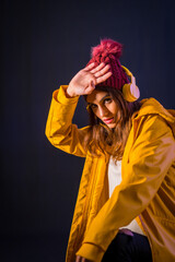 Portrait of a young brunette Caucasian with headphones, yellow jacket and wool hat on a black background, pretty girl listening to music looking at camera