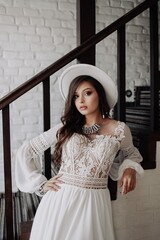 A beautiful brunette bride with long hair stands in a house near a wooden staircase and a white brick wall in a white dress, white hat, and jewelry