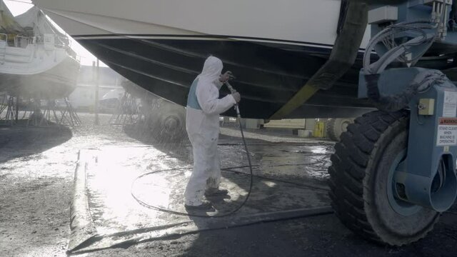 Male hourly staff worker in full suit for skilled labor job uses air compressor to blast spray power wash underside bottom of boat on lift at marina for season to winterize vessel for owner