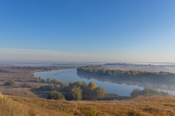 Autumn landscape in the early morning overlooking the river. A wide river and endless expanses of fields. Yellow leaves on trees and bushes are illuminated by the rays of the rising sun.