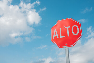 Close-up of a red Stop sign in Spanish right aligned against the blue cloudy sky. Red Alto, background with space for text. - 476922566
