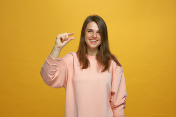 Smiling young girl showing little size gesture with fingers on yellow studio background. Small prices, big discounts
