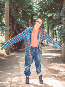 A girl with two braids in a plaid shirt with overly long sleeves and huge jeans stands spreading her arms to the side in the park on a green alley. Post-processing. Artistic effect