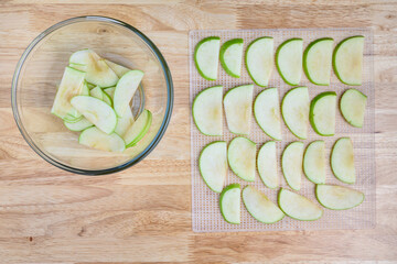 Glass bowl full of granny smith apple slices on a butcher block table, and slices laid out on a mesh tray ready for dehydrating apples
