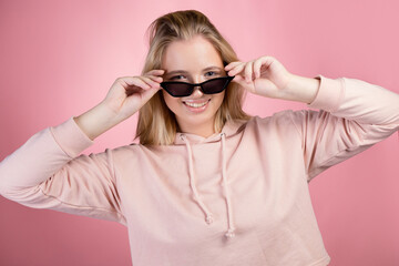 Stylish young and trendy blonde in a pink oversized hoodie and black sunglasses, portrait on a pink background