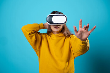 VR gamer, a young woman in a youth outfit uses a virtual reality helmet. Brunette in a yellow hoodie on a blue background with a virtual reality helmet on her head, touching something in 3d