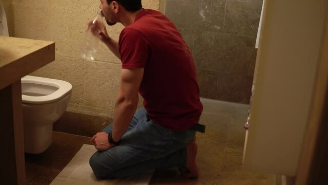 A sick man is sitting in front of toilet and drinking pure water from the bottle after vomiting. He looks sick and tired. Young caucasian guy feels stomachache and nausea. Consequences of intoxication