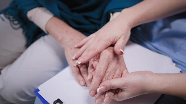 support hands close up, female therapist in blue uniform with clipboard holds elderly woman patient hands during medical visit