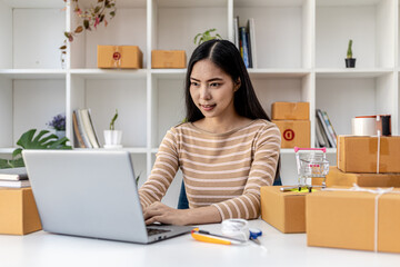 A beautiful Asian business owner opens an online store, she is checking orders from customers via laptop, sending goods through a courier company, concept of a woman opening an online business.