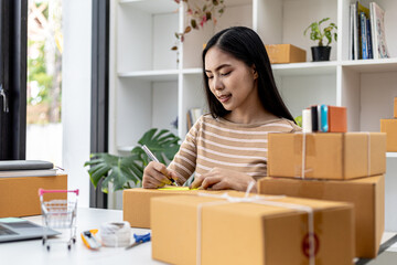 Asian woman writing customer's shipping information on parcel box, she owns an online store, she ships products to customers through a private courier company. Online selling concept.