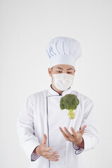 Restaurant chef in medical mask tossing broccoli, isolated on white